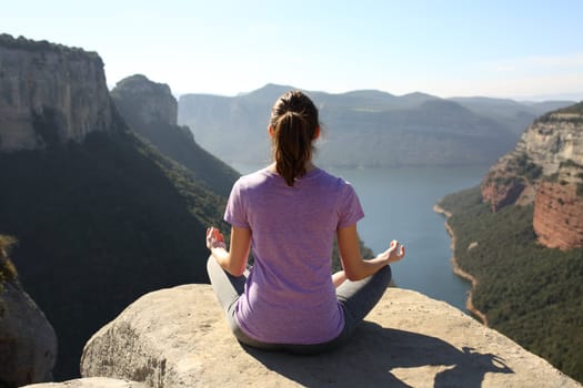 Back view of a yogi practicing yoga on the top of a cliff in the mountain