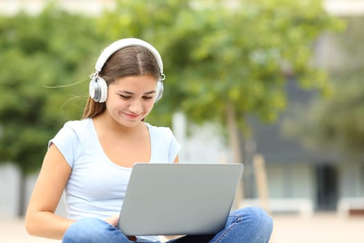 Happy student elearning using laptop and headphones sitting in a campus