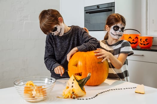 Two young children with skull facepaint cutting a pumpkin in their kitchen.