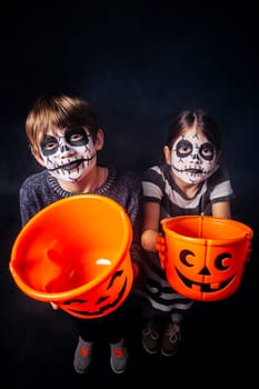 Two children with skull makeup holding pumpkin pails at Halloween.