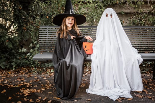 Young boy dressed in a bed sheet to be a ghost, and a young girl in witch costume holding a pumpkin pail for halloween.