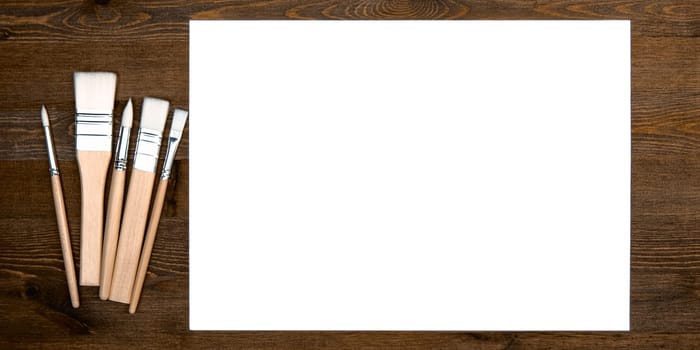 A clean white sheet and brushes on a wood textured background with space to copy. Layout, mockup space for text and drawing.