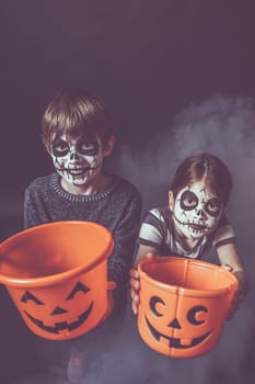 Two children with skull makeup holding pumpkin pails at Halloween.