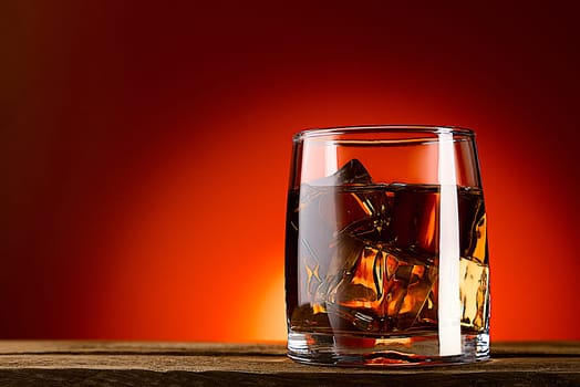A glass of whiskey or cognac and ice cubes, close-up on a wooden table. Red background with gradient. Space for labels, text and logo. Layout for advertising.