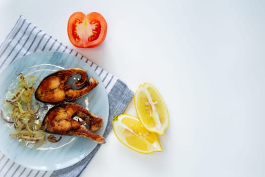Fried fish with onions lies on a plate, on a waffle towel, next to a lemon and a tomato on a white background. High quality photo