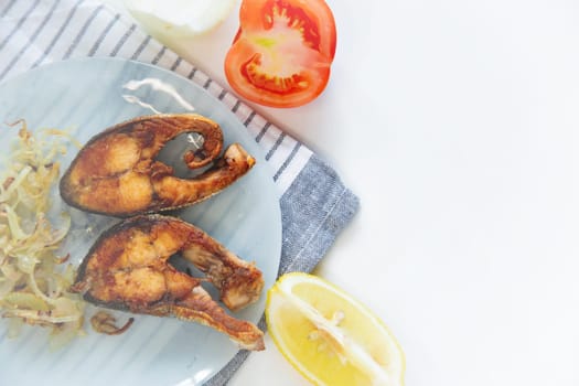 Fried fish with onions lies on a plate, on a waffle towel, next to a lemon and a tomato on a white background. High quality photo
