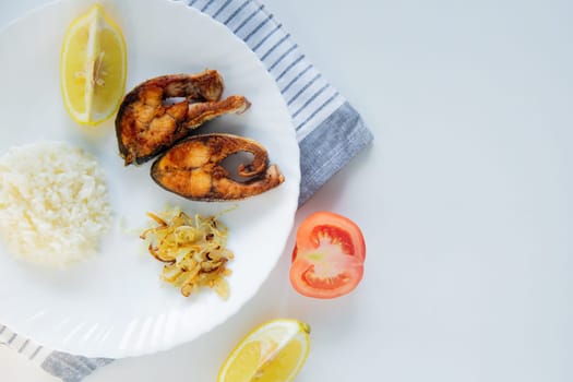 Fried fish with onions, rice and lemon lies on a plate, on a waffle towel and cutlery on a white background. High quality photo
