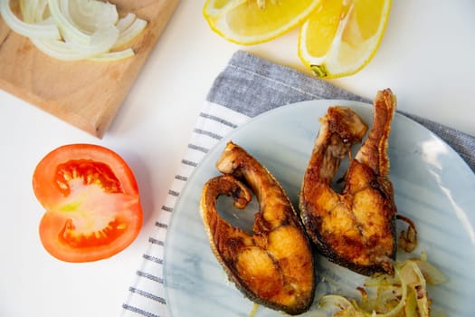 Fried fish with onions is on a plate, on a waffle towel, next to it is a cutting board with chopped onions, lemon and tomato on a white background. High quality photo