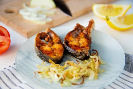 Fried fish with onions, rice and lemon is on a plate, on a waffle towel, next to it is a cutting board with chopped onions on a white background. . High quality photo