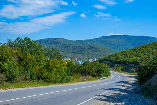 The road in the mountains, where green trees grow, goes around a bend, in the distance you can see a city in the lowlands. High quality photo