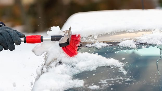 Human hand cleaning car from snow in wintertime day. Person in gloves with removing snowflakes from windshield in close-up. Brushing with broom on white automobile.