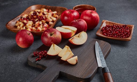 Still life with fresh lingonberries, red appples with knife on black cutting board and  homemade jam on wooden bowl on dark background. Top view. Autumn harvest, Home canning.