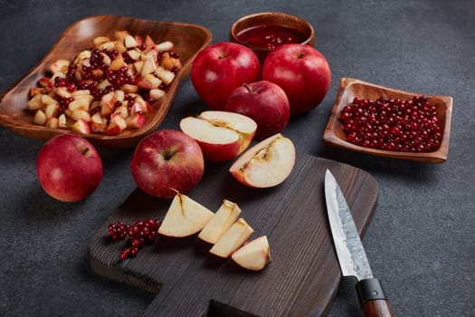 Still life with fresh lingonberries, red appples with knife on black cutting board and  homemade jam on wooden bowl on dark background. Top view. Autumn harvest, Home canning.