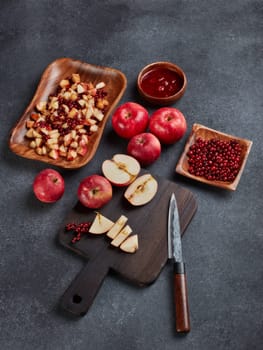 Still life with fresh lingonberries, red appples with knife on black cutting board and  homemade jam on wooden bowl on dark background. Top view. Autumn harvest, Home canning.