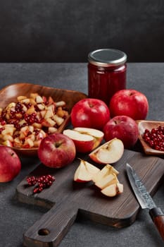 Still life with fresh lingonberries, red appples with knife on black cutting board and jar of homemade jam on dark background. Autumn harvest, Home canning.