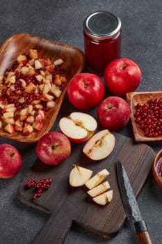 Still life with fresh lingonberries, red appples with knife on black cutting board and  homemade jam on wooden bowl on dark background. Top view. Autumn harvest, Home canning.