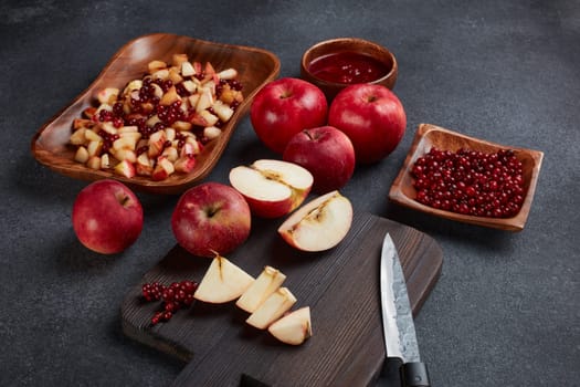 Still life with fresh lingonberries, red appples with knife on black cutting board and  homemade jam on wooden bowl on dark background. Top view. Autumn harvest, Home canning.