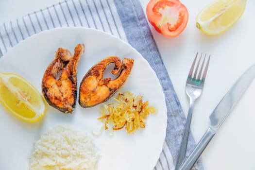 Fried fish with onions, rice and lemon lies on a plate, on a waffle towel and cutlery on a white background. High quality photo
