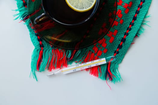 A cup of tea with lemon stands on a dark saucer, which stands on a knitted green-red scarf, next to it lies a thermometer on a white background. . High quality photo