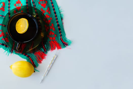 A cup of tea with lemon stands on a dark saucer, which stands on a knitted green-red scarf, next to it lies a thermometer and a lemon on a white background. High quality photo