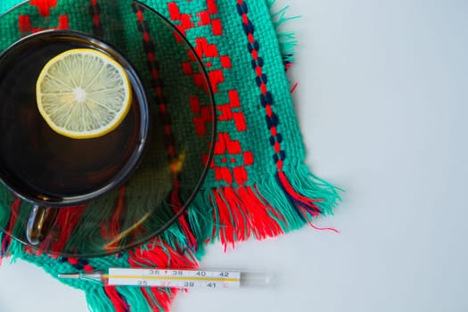A cup of tea with lemon stands on a dark saucer, which stands on a knitted green-red scarf, next to it lies a thermometer on a white background. . High quality photo