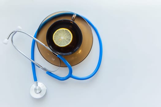 The blue stethoscope lies on a white background with a mug of tea on a saucer with a lemon in the middle. High quality photo