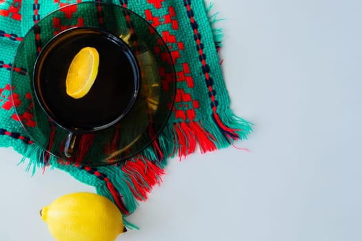 A cup of tea with lemon stands on a dark saucer, which stands on a knitted green-red scarf, next to it lies a thermometer and a lemon on a white background. High quality photo