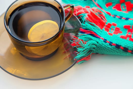 A dark transparent cup of tea with lemon stands on a dark saucer, next to it lies a knitted winter scarf, on a white background. High quality photo