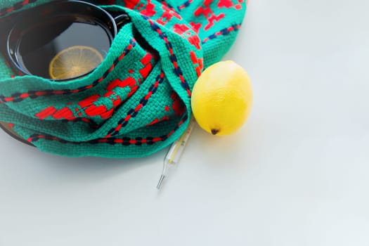 A cup of tea with lemon stands on a knitted green and red scarf, there is a thermometer and a lemon next to it. High quality photo