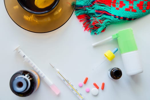 A cup of tea with lemon stands on a dark saucer, a knitted winter scarf lies next to it, medicine, pills, a thermometer, medicine and drops from a cold on a white background lie next to it in a circle. High quality photo