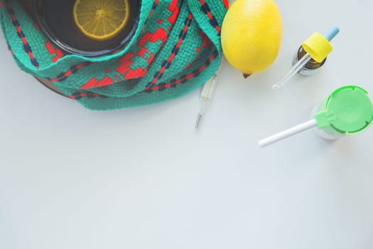 A cup of tea with lemon stands on a knitted green and red scarf, next to it is a thermometer, lemon and drops from a runny nose and a throat spray. High quality photo