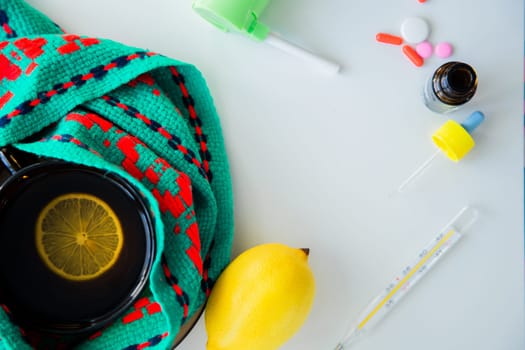 A cup of tea with lemon stands on a knitted green and red scarf, next to it is a thermometer, lemon and drops from a runny nose and a throat spray. High quality photo