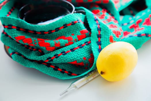 A cup of tea with lemon stands on a knitted green and red scarf, there is a thermometer and a lemon next to it. High quality photo