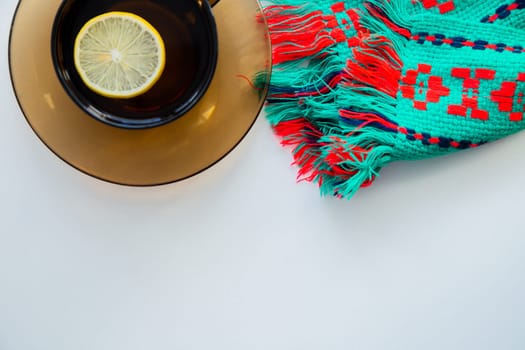 A dark transparent cup of tea with lemon stands on a dark saucer, next to it lies a knitted winter scarf, on a white background. High quality photo