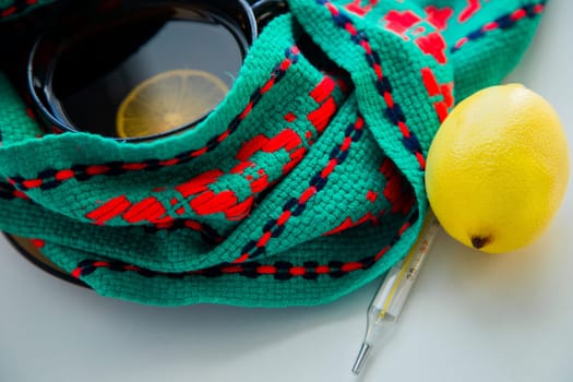 A cup of tea with lemon stands on a knitted green and red scarf, there is a thermometer and a lemon next to it. High quality photo