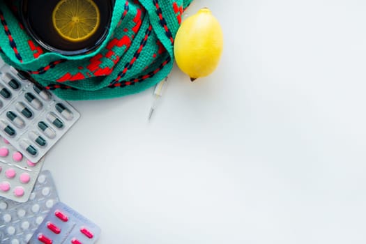 A cup of tea with lemon stands on a knitted green and red scarf, there is a thermometer next to it, a lemon and pills on the side. . High quality photo