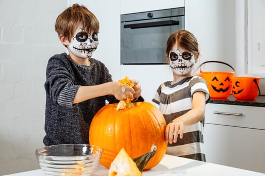 Two young children with skull face paint carving a pumpkin in their kitchen.