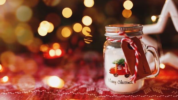 Festive Christmas candle in form of glass cup or mug and other decorations, blurred background with Christmas fir tree lights, garlands and candles. Red ornament on tablecloth. Festive mood at home