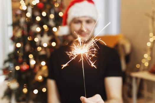 Smiling man in red santa hat or cap holding burning Christmas or New Year sparkler lights in hand. Christmas fir tree with garland lights on blurred background. Festive mood. High quality photo