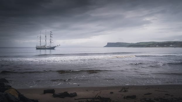 Dark, dramatic storm sky rolling over anchored tall ship near Northern Ireland coast with Fair Head in far distance, Ballycastle