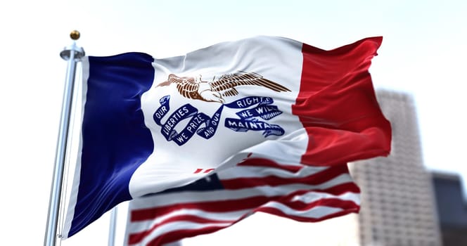 the flag of the US state of Iowa waving in the wind with the American stars and stripes flag blurred in the background. Three vertical stripes. The center stripe contains an eagle holding a ribbon.