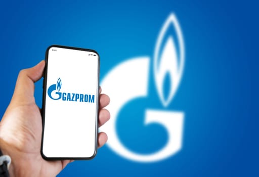 Saint Petersburg , RUS, October 2021: hand holding a phone with the Gazprom mobile app on the screen and the logo blurred on a blue background. Gazprom is a Russian multinational energy corporation