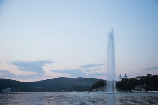 A fountain several meters high spouts from the center of the lake, you can see the mountains and the church, evening. Abrau Durso, Krasnodar Territory. High quality photo
