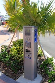 LAUDERDALE-BY-THE-SEA, FL, USA - APRIL 7, 2014: Closeup of an electric parking meter with signage in the shade of palm trees and plants of a newly redesigned parking lot. New silver gray parking meter with digital display panels in a shady area of a newer walkway and parking lot buildings and a few people on the sidewalk.