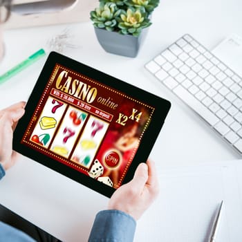 Online gambler gambling at the office holding his tablet computer showing a casino interface with lucky numbers above his desk and workspace