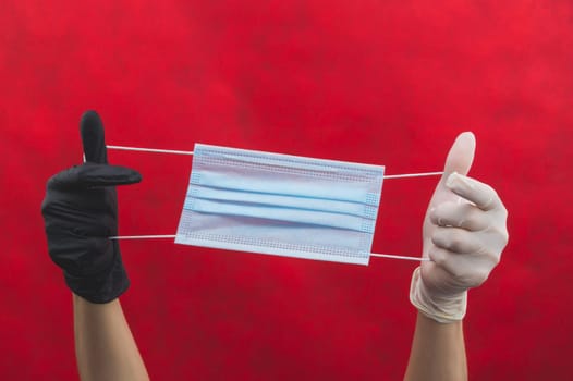 Hands in different protective gloves stretch a medical mask on a red background. one hand in black glove, the other in white. preventive measures against respiratory illness and virus