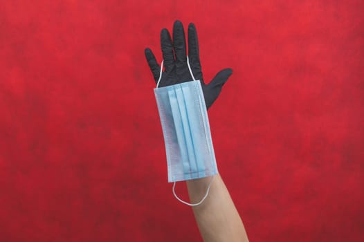 Blue medical mask hangs on the hand with a black surgical glove. preventive measures against respiratory illness and virus. red background with copy space