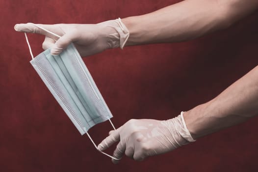 Hands in protective gloves stretch a medical mask on a red background. preventive measures against respiratory illness and virus. hands in white surgical gloves holds disposable mask for protection