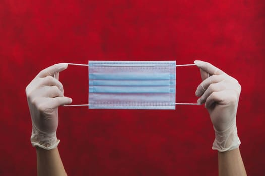 Blue medical mask in hands with white gloves on a red background. preventive measures against respiratory illness and virus. hands in white surgical gloves holds disposable mask for protection