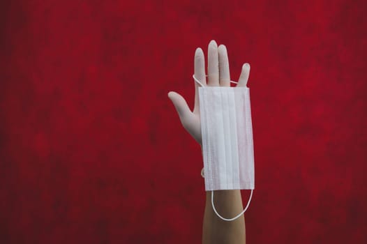 White medical mask hangs on the hand with a white surgical glove. preventive measures against respiratory illness and virus. red background with copy space